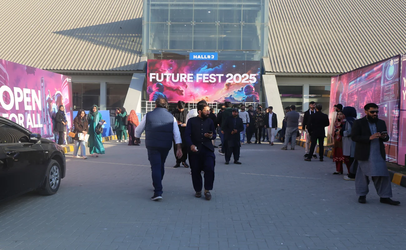 A diverse group of people strolls past a large screen promoting Future Fest, showcasing excitement and anticipation for the event.